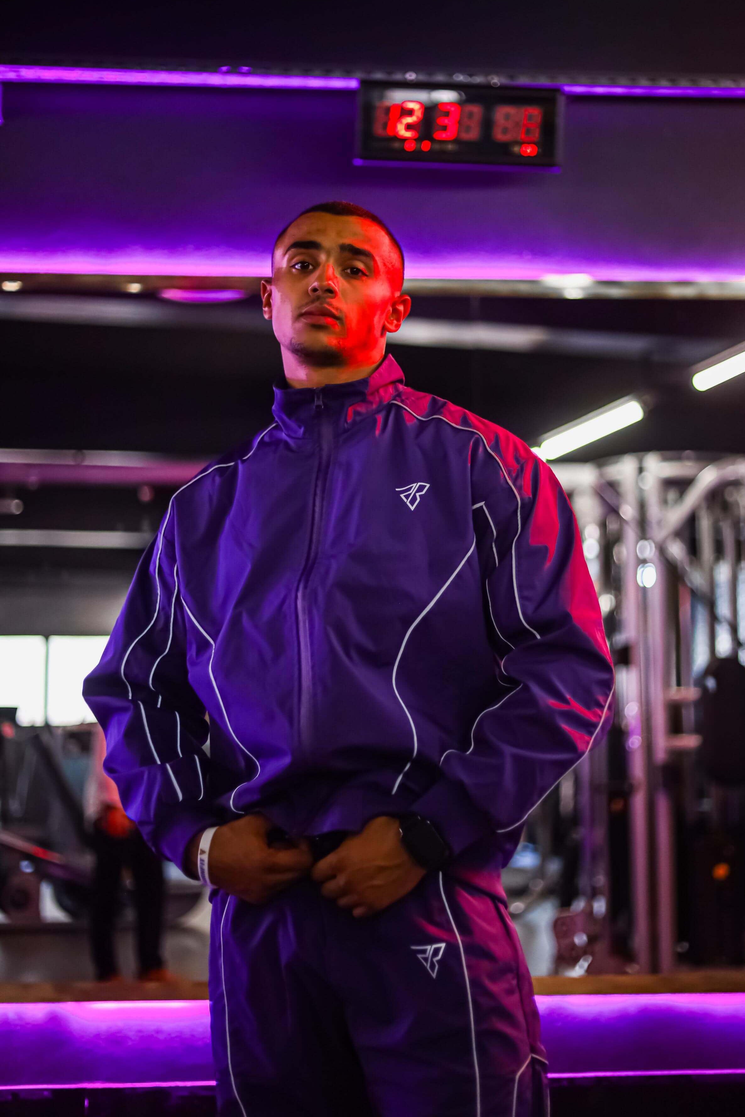 Person wearing a purple tracksuit in a gym setting with digital clock displaying time.